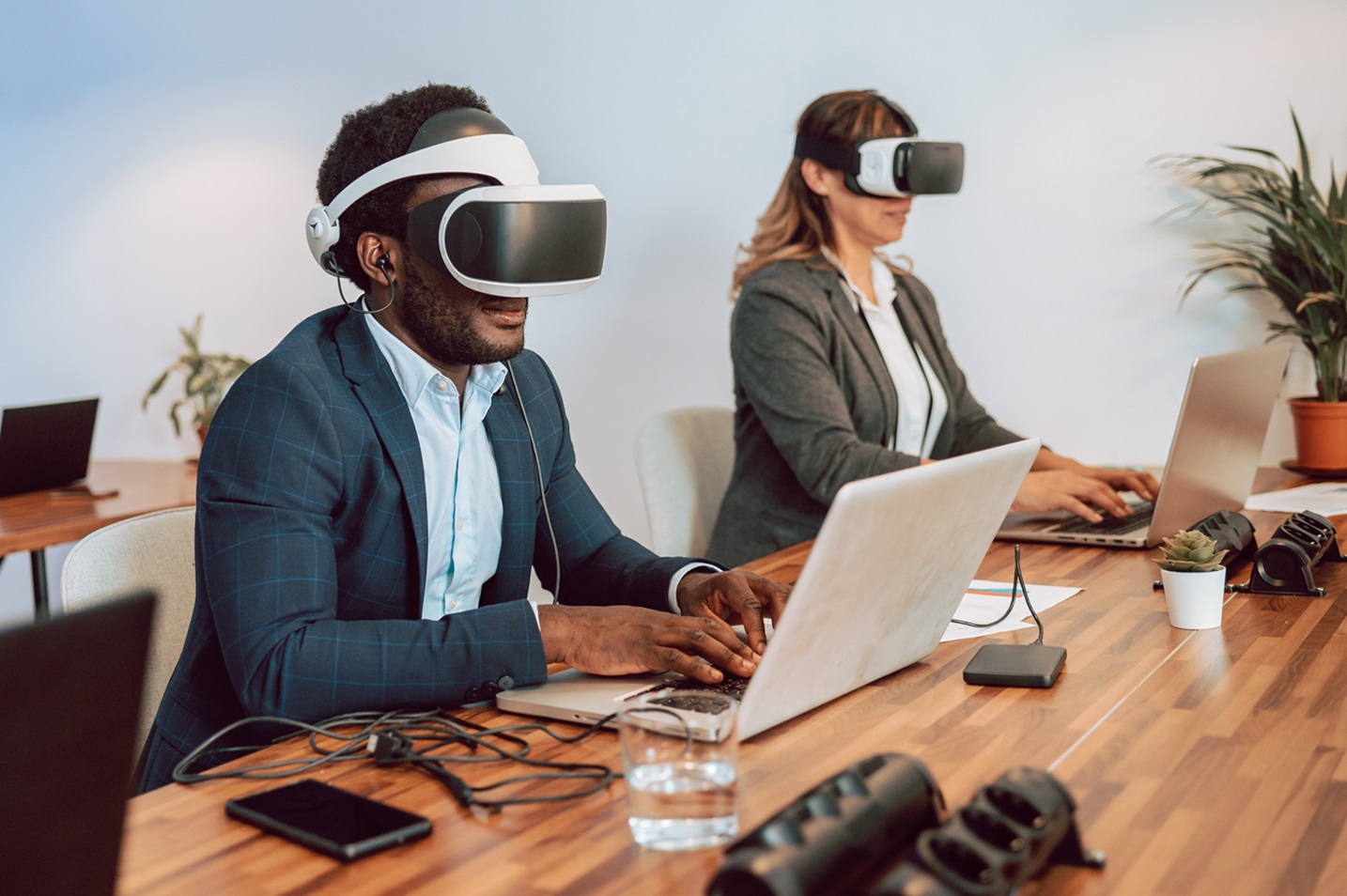 Concentrated African American male entrepreneur in VR goggles sitting at table with laptop and working on project while working in office